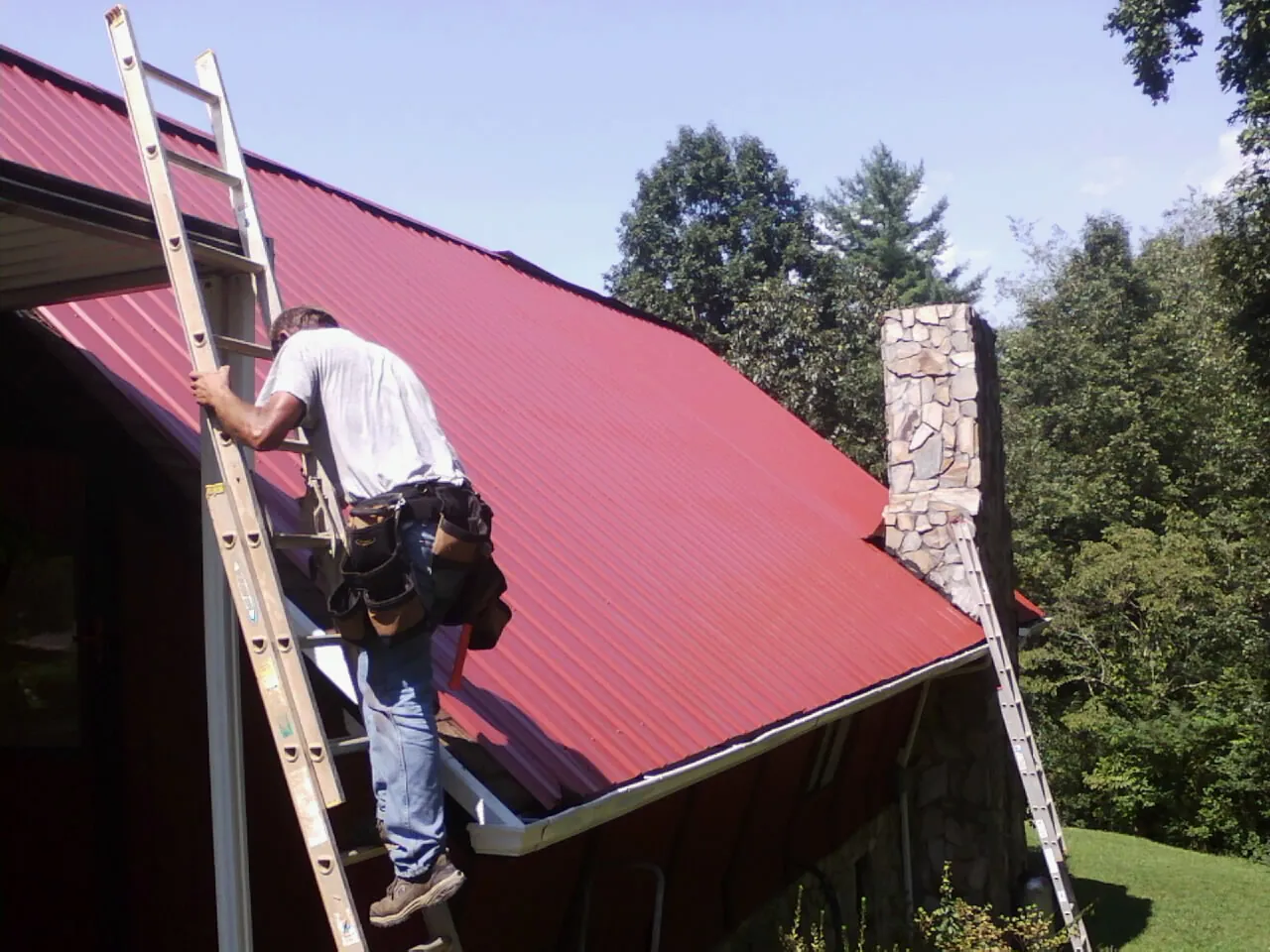 Roofer on a ladder beside a red metal roof and stone chimney — Soffit & Fascia Repair by Roofing Brothers in Careywood